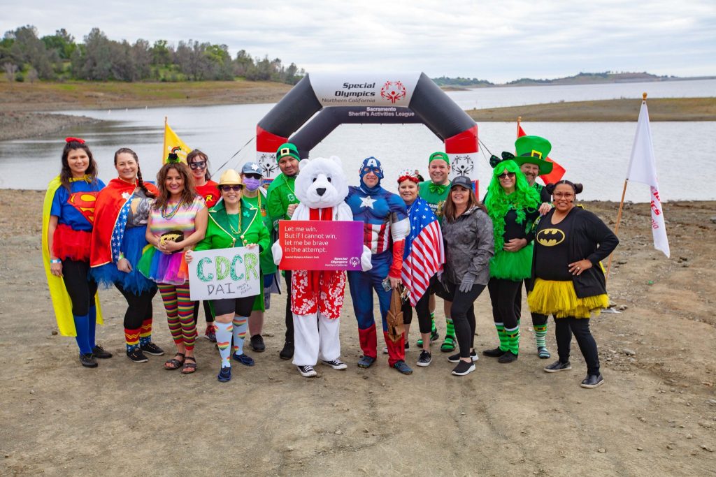 A group pf people in various superhero and leprechaun costumes stand in front of Folsom Lake