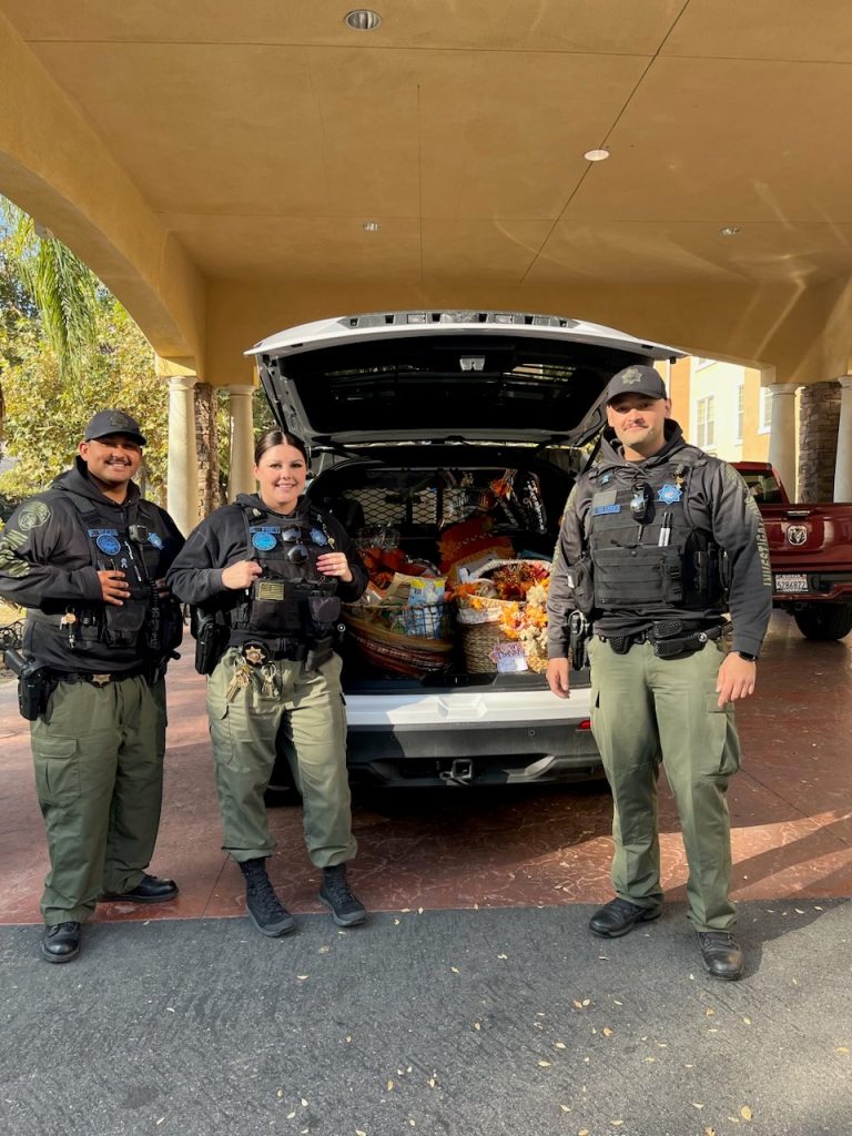 Three officers in uniform stand by the open trunk of a vehicle full of Thanksgiving baskets