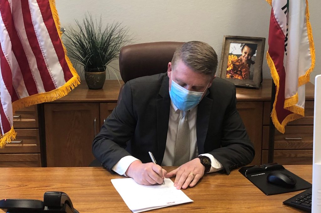 Man in a suit and a mask signs a document at a desk