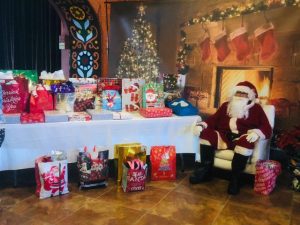 Santa Claus sits next to a table piled with Christmas gifts.