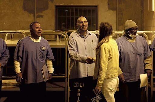 An incarcerated person shakes hands with a woman in a prison housing unit while other incarcerated people look on.