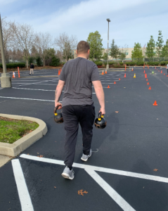 A man with back to the camera holds kettlebells and walks a course lined with cones.
