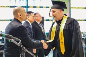 A man in a graduation cap and gown shakes hands with a man in a suit.