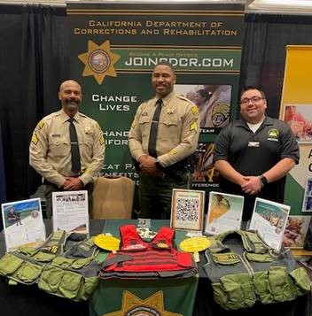 Two men in correctional officer uniforms and a man in a black polo shirt stand behind a table covered in CDCR information and gear.