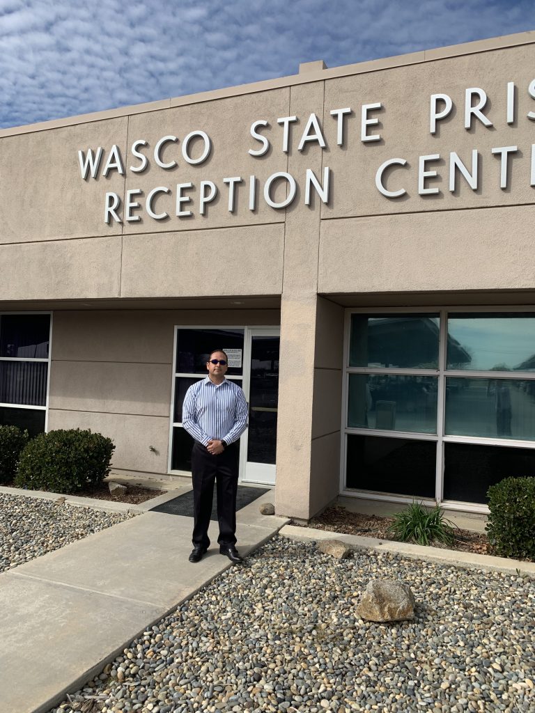 A man in a striped shirt stands in front of a building with a sign: "Wasco State Prison Reception Center."