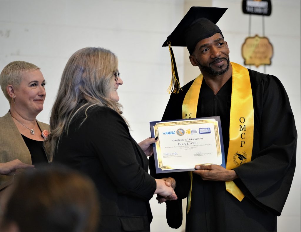 A man in a cap and gown shakes hands with a woman handing him a diploma.