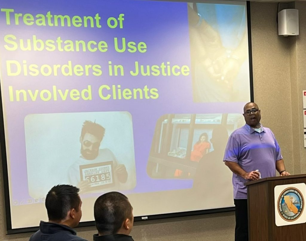 A man stands at a lectern in front of a presentation titled "Treatment of Substance Use DIsorders in Justice Involved Clients"