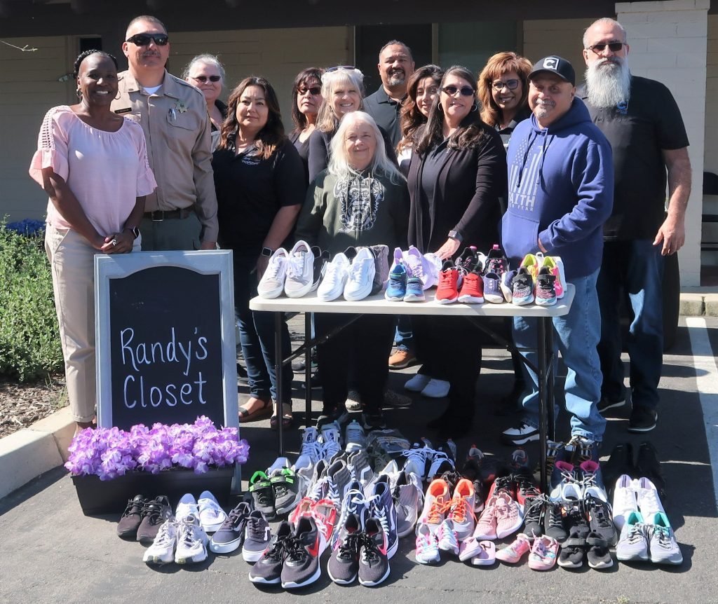 A large group of people stand at a table. The table and ground are full of shoes. A chalkboard sign says "Randy's Closet."