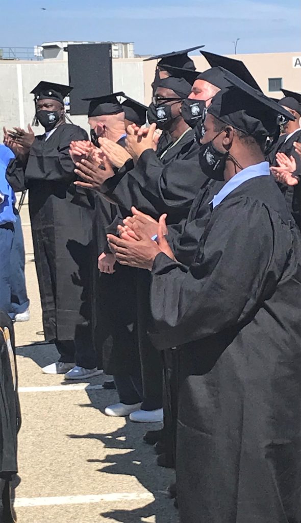 Students in caps and gowns applaud at an outdoor graduation ceremony.