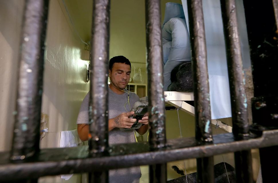 A man wearing headphones looks at a tablet in a prison cell.