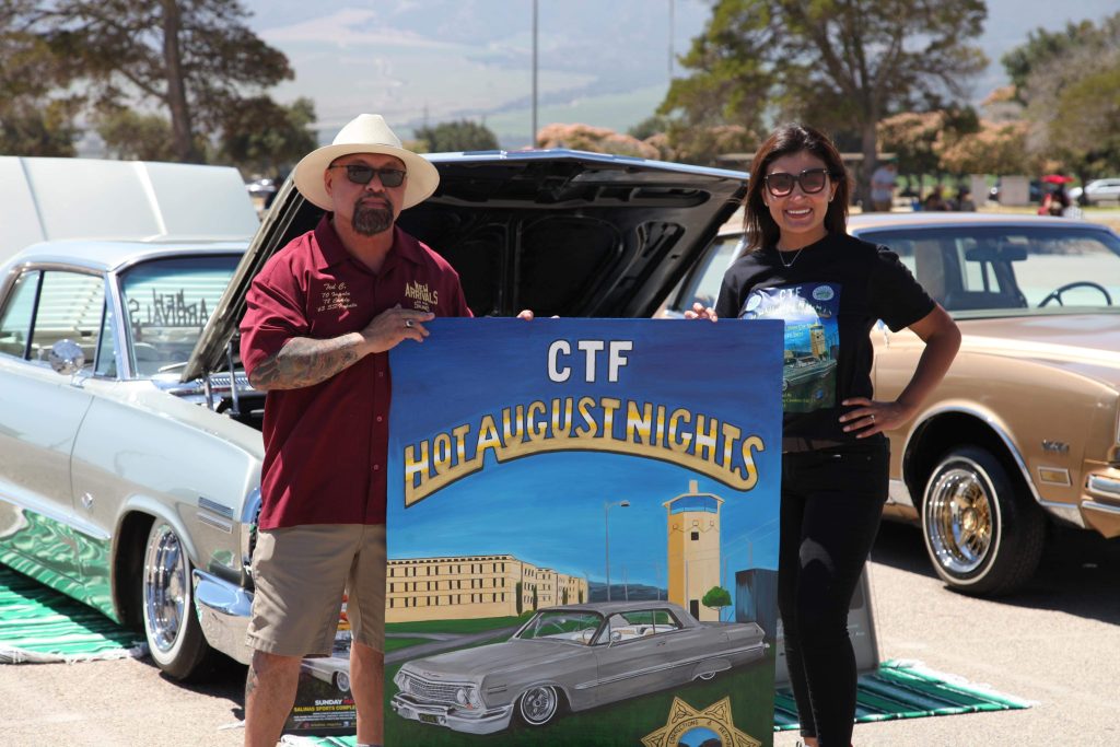 two people standing in front of cars with a sign for CTF Hot August nights car show