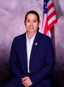 A woman in a dark jacket stands in front of an American flag.