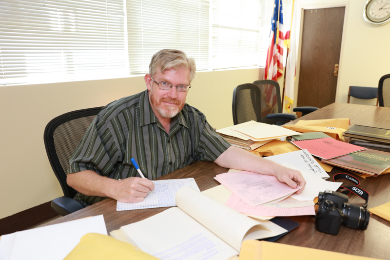 A man with blonde hair sits at a desk covered with papers and a camera.