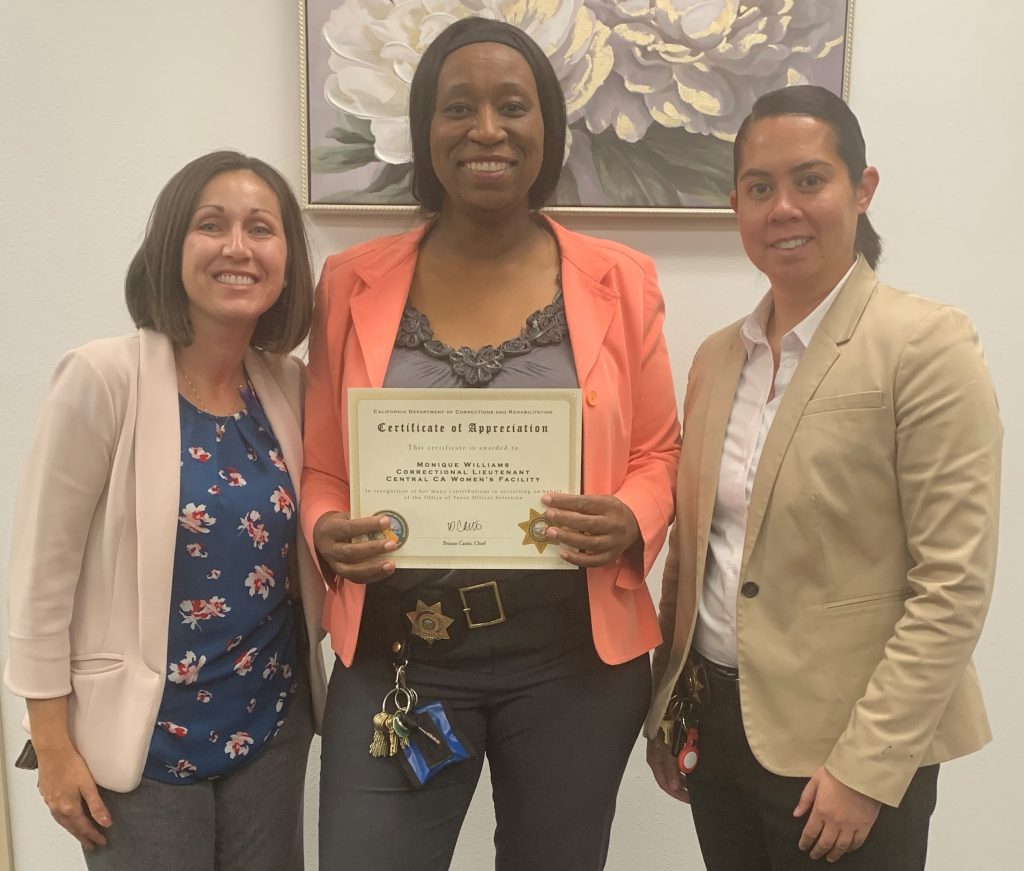 Three women pose for a photo, the woman in the middle holds a certificate of recognition