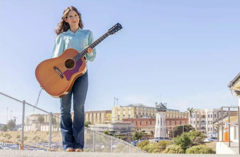 A woman holding a guitar stands outside a prison