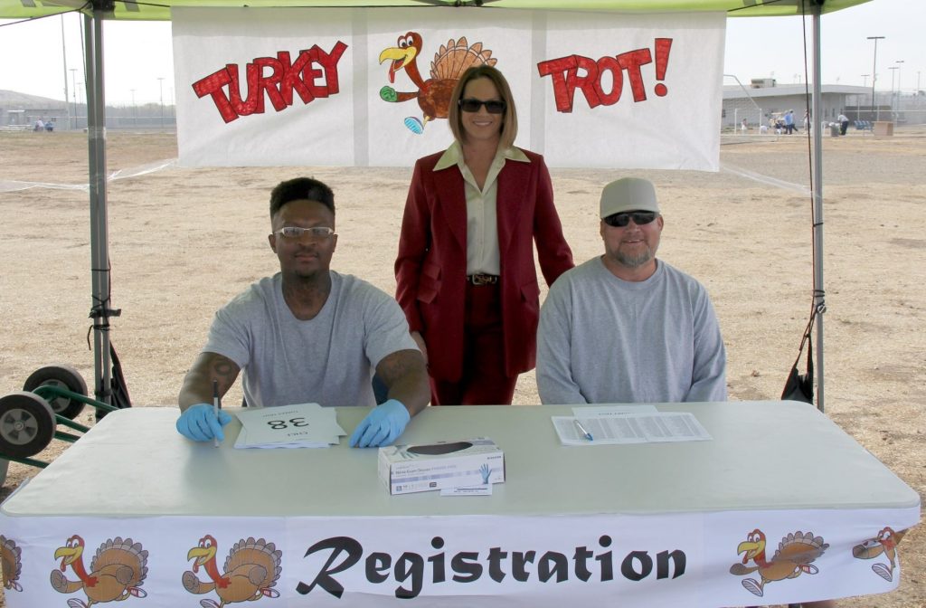 Two men sit at a table under a sign that says "Turkey Trot." A woman in a red pantsuit stands between them.